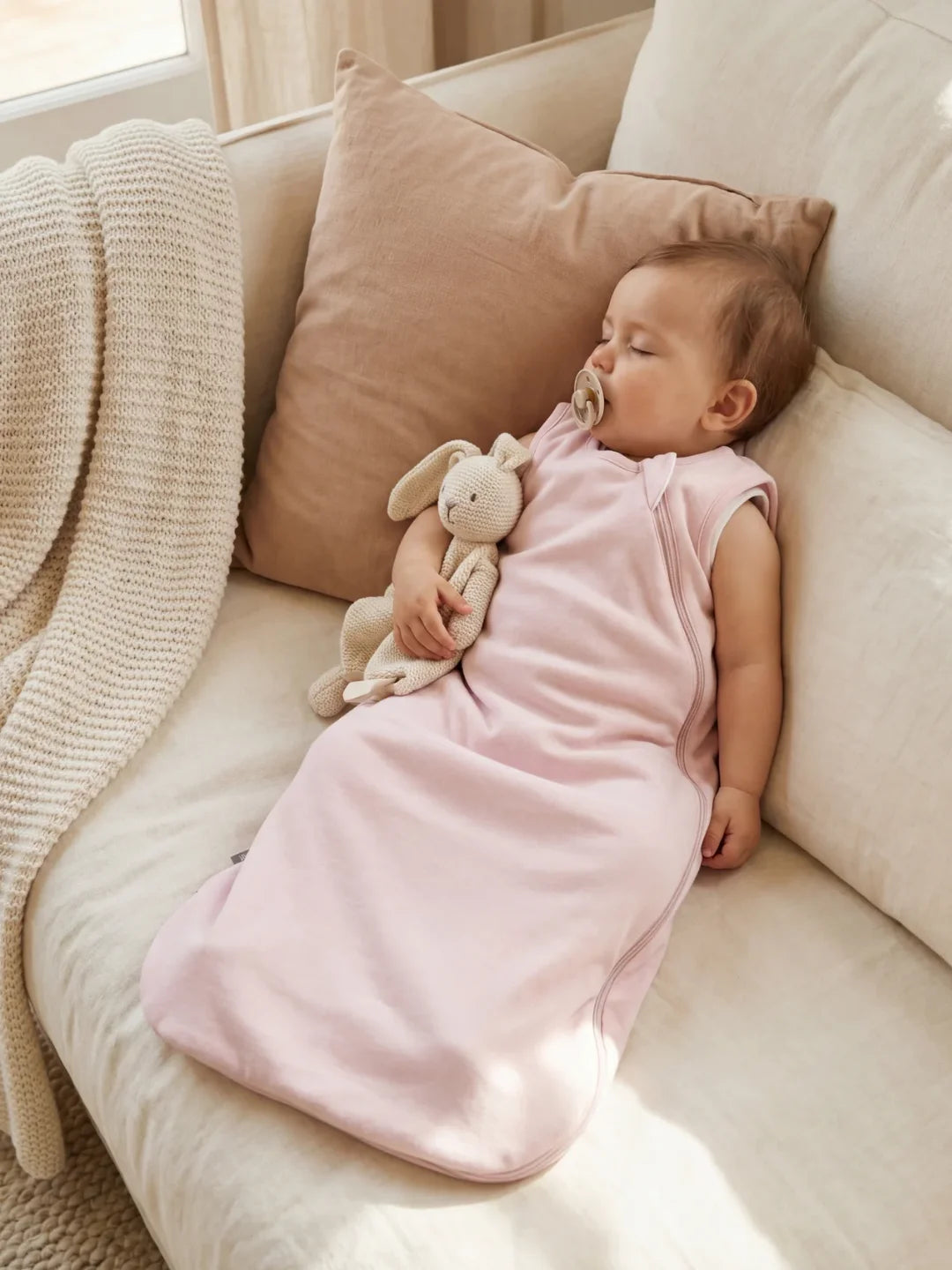 Bébé endormi dans sa gigoteuse jersey rose pâle avec doudou lapin sur canapé