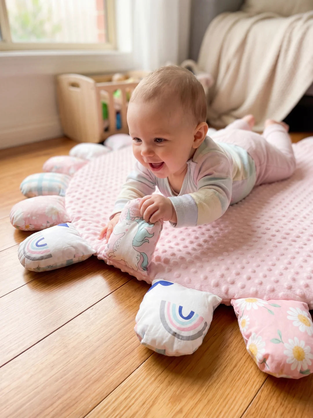 Bébé allongé sur le ventre sur un tapis rose, attrapant un pétale illustré d'une licorne pour développer sa motricité.
