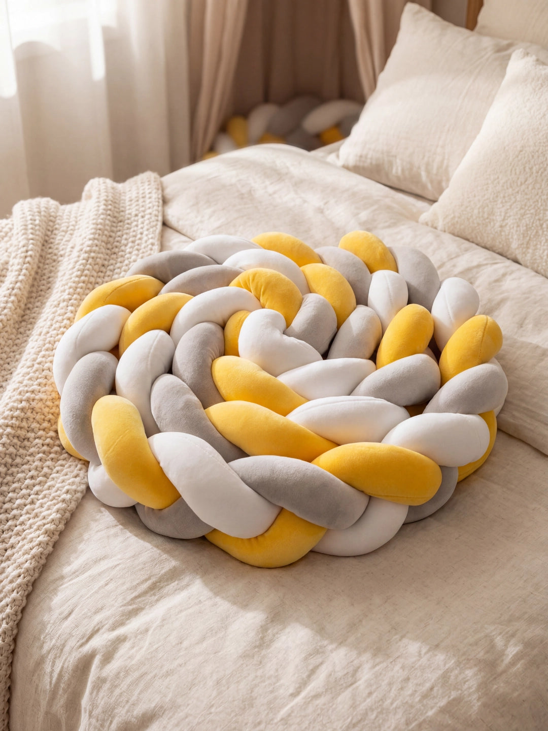 Gros plan sur un coussin nœud tressé rond aux couleurs scandinaves (jaune, gris, blanc) posé sur un lit.
