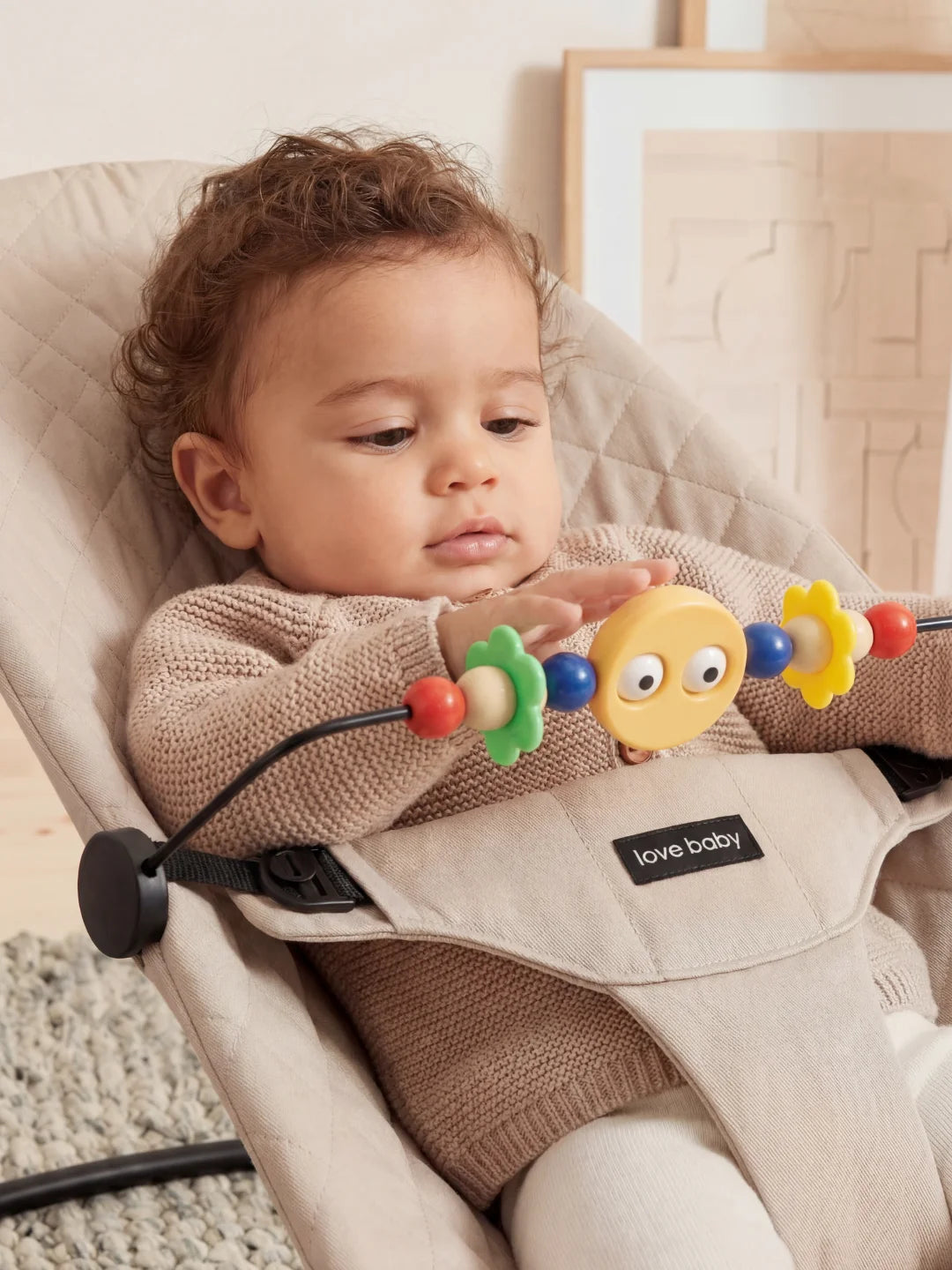 Gros plan d'un bébé curieux jouant avec les fleurs colorées d'une arche d'éveil fixée sur son transat beige.