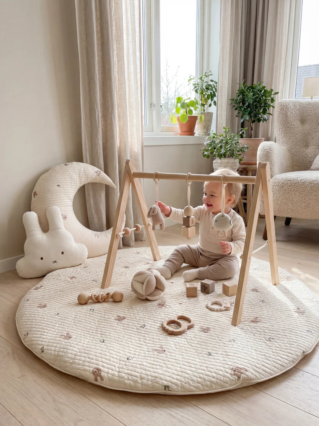 Bébé assis sous un portique d'éveil en bois sur un tapis rond à motifs lapins, avec des accessoires de jeu suspendus.