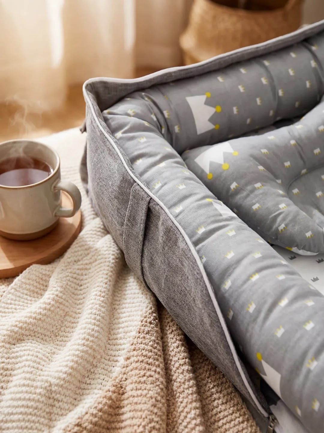 Réducteur de lit gris avec motif couronne, posé sur couverture beige, tasse de thé à côté.
