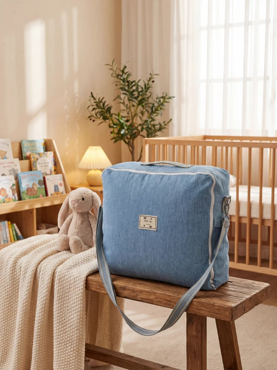Sac bleu posé sur un banc en bois dans une chambre de bébé avec une peluche lapin et un lit à barreaux en arrière-plan.