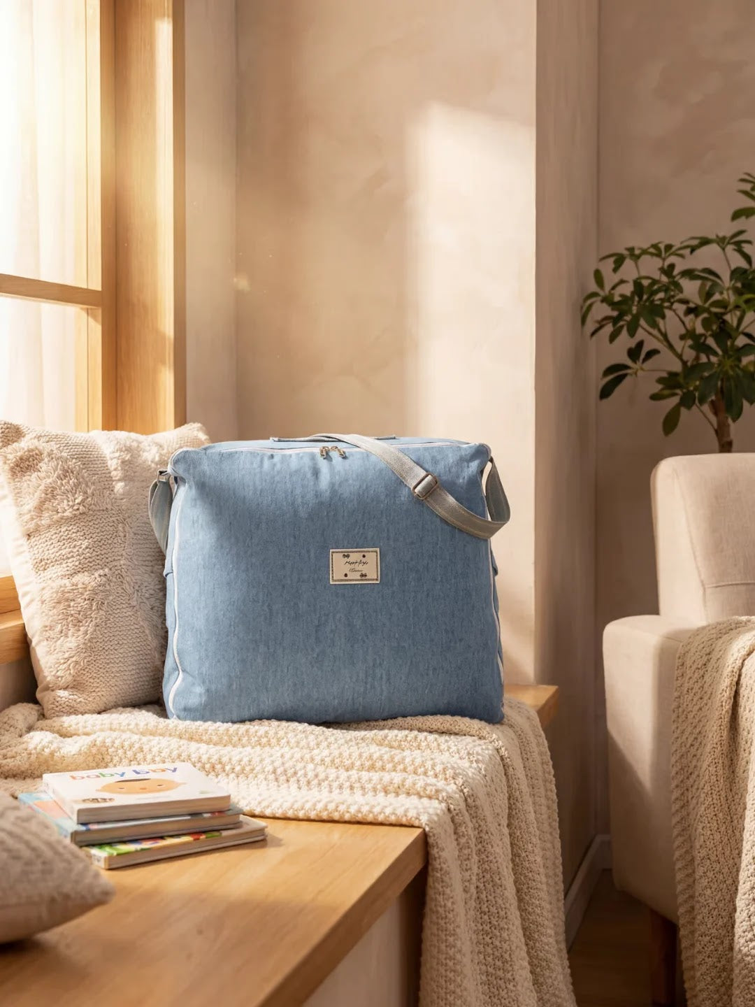 Sac bleu clair posé sur un banc près d'une fenêtre dans un intérieur cosy avec coussins et plante.