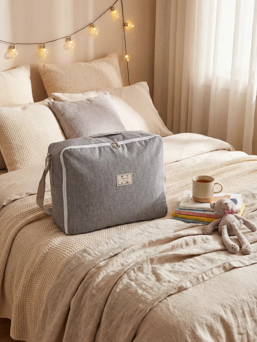 Sac de rangement gris sur un lit beige avec livres, tasse et peluche, guirlande lumineuse en arrière-plan.
