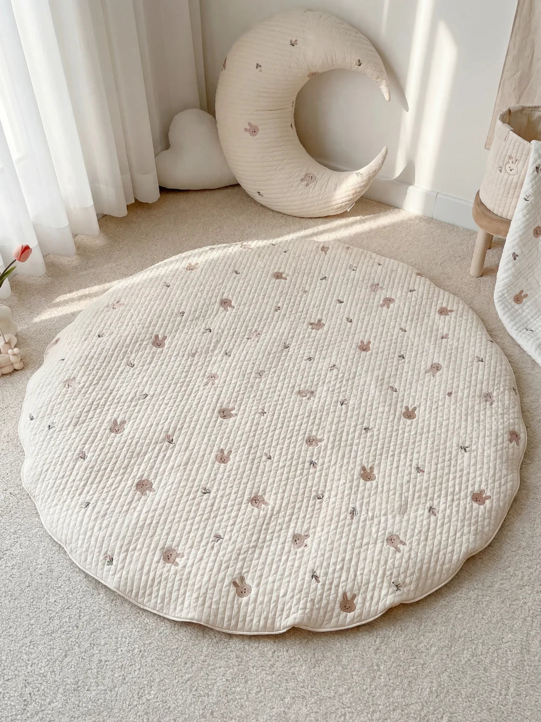 Grand tapis d'éveil rond blanc avec motifs de têtes de lapins beiges, posé dans un coin lecture avec un coussin lune.