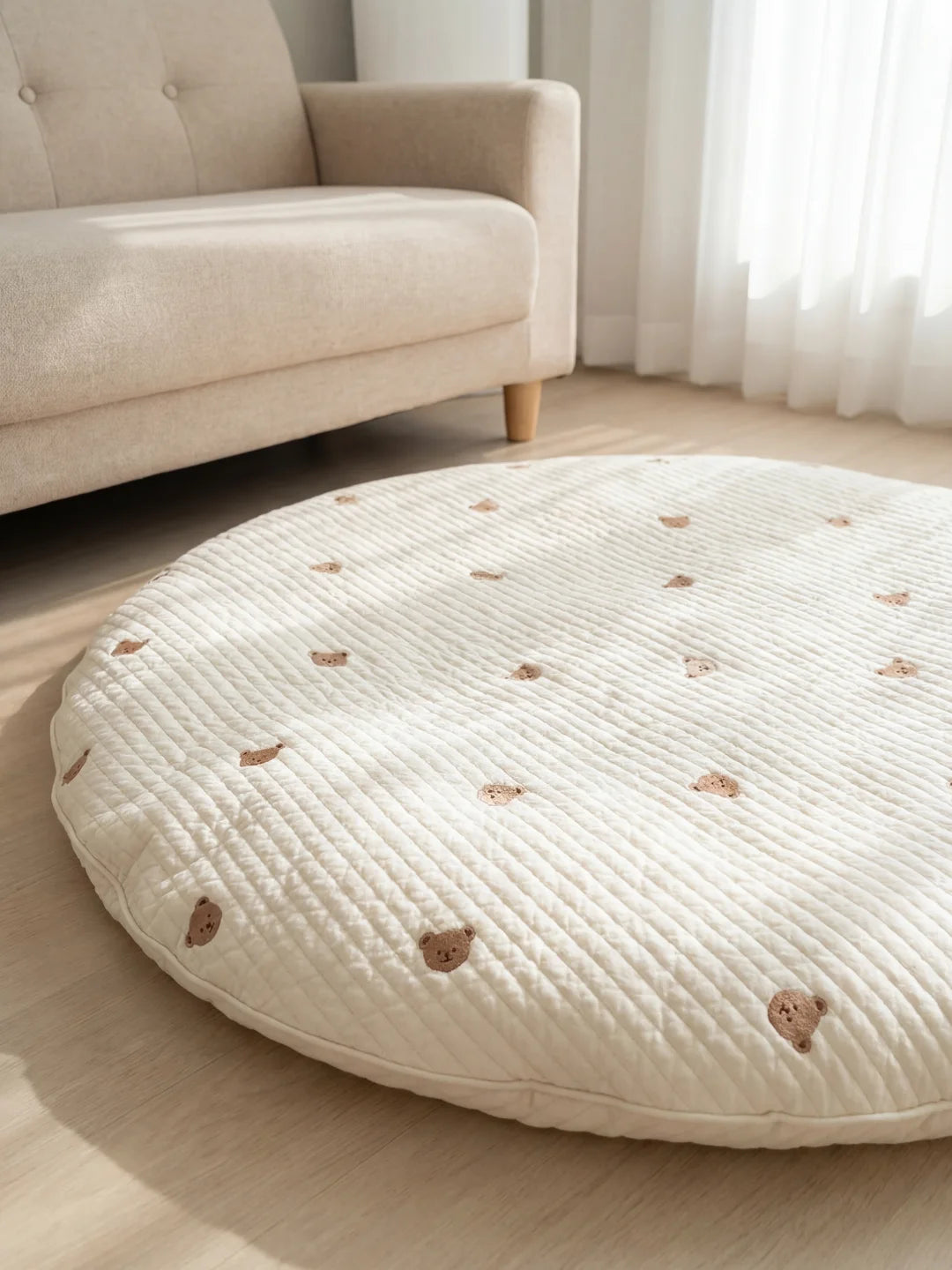 Vue d'ensemble d'un tapis de sol matelassé pour bébé de couleur crème avec motifs ours, placé devant un canapé beige.