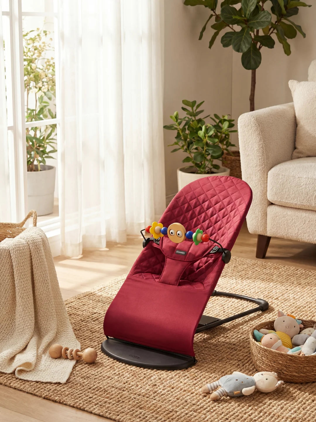 Transat pour nouveau-né couleur bordeaux avec arche de jeux, posé sur un tapis en jute dans un intérieur moderne.