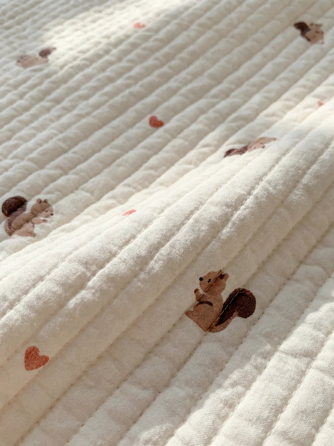 Gros plan sur la texture du tissu matelassé blanc avec broderies d'écureuils et petits cœurs terracotta.